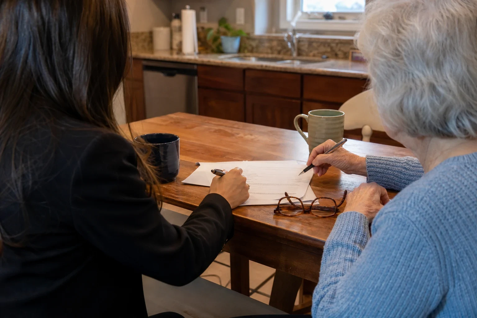 An agent and an older woman with gray hair sit at a wooden kitchen table with papers, pens, coffee mugs and reading glasses, a kitchen sink and window behind them