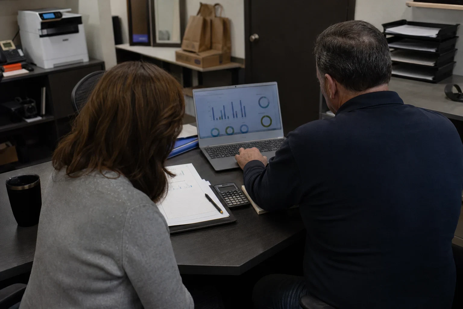 Two people seen from behind at a dark office desk reviewing bar and donut charts on a laptop, with printed reports, a calculator and pen beside them in a small back office